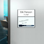 Rumsskylt med whiteboardtavla med tillbehörande penna utanför ett kontor